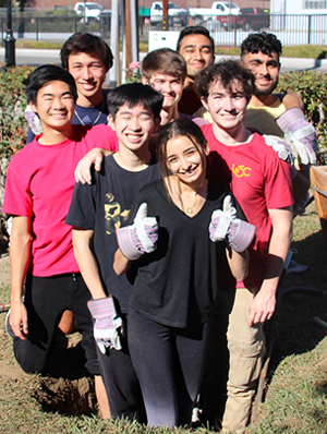 A group of students standing and posing in a school garden, smiling from the camera. In the front a young girl smiles, her thumbs up, her hands in gardening gloves.