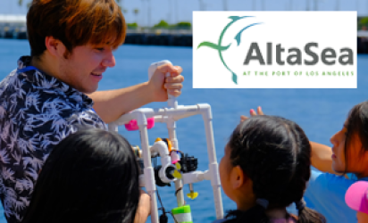 An instructor in a Hawaiian style blue and white shirt, holding up a PVC pipe contraption with neon pink and green additions, standing in front of a group of young girls, an out of focus shot of the ocean, a far shore, and palm trees in the background. The AltaSea Logo is on white over the top right corner of the photo.