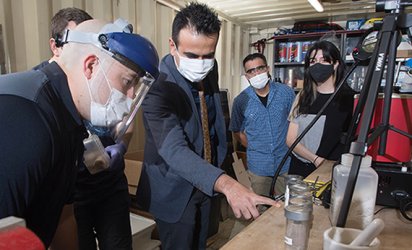 A group in masks gathered around samples from a thermal energy storage experiment. The man in the center wears a suit and points at the samples. The man on the left (in a face mask) leans over and examines the samples. Two more individuals stand in the background in front of a storage shelf watching. One individual is obscured behind the individual on the left. Various scientific equipment near the right edge of the photograph.