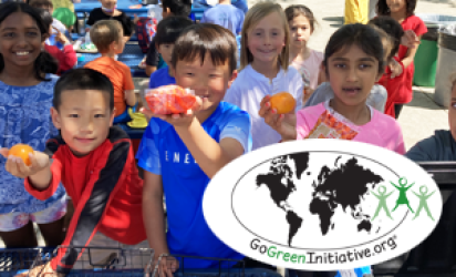 Children holding up fruits towards the unseen camera, other children behind them in the background. The GoGreen initiative logo is on a white oval over the bottom right corner of the image.