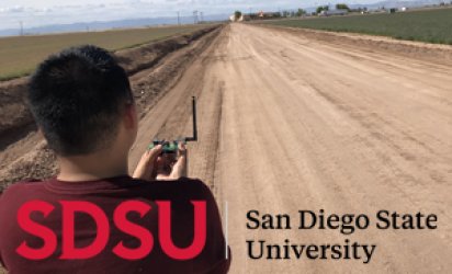 The back of a Smart Grid Lab student's head on the left of the frame, a dirt road stretching towards the horizon in the center of the shot, with farmland stretching on either side, disappearing into a slanted horizon line against blue skies and clouds. The Sand Diego State University logo runs across the bottom of the shot, a red SDSU over the back of the student's shirt, and San Diego State University spelled out in black over the dirt road.