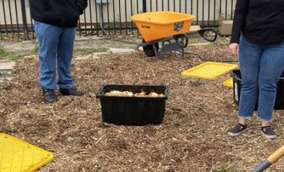 Two youth on either end of the image raking up unseen compost. The midground features mulch with bins of compost, and adults (somewhat obscured) behind the youth.