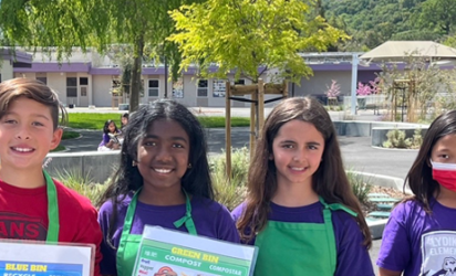 Seven kids standing almost shoulder to shoulder, many wearing green aprons. The two in the center are holding up laminated composting and recycling infosheets. A green space, and school buildings are in the background.