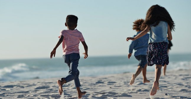 Three kids, their backs to the viewer, run across the sands of the beach, waves lapping against the shore out of focus in the background.