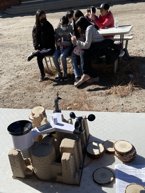 Students sit at a table at the Skyland Ranch in the background, a weather station in the foreground.
