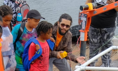 Students and instructors gathered on a dock looking at a contraption built of PVC pipe ging lowered by an orange crane arm into the water off the dock Seaweed or kelp hangs off hooks attacked to the contraption.