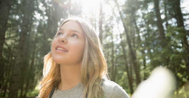 A young woman smiling, surrounded by trees, the sun shining through the trunks behind her.